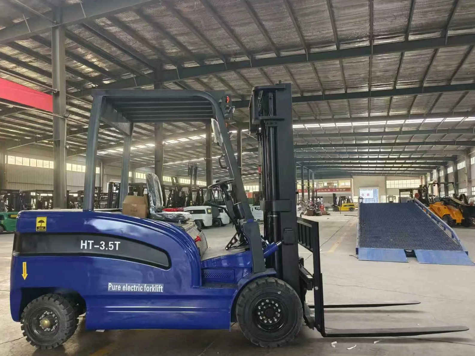Side view of a blue electric forklift inside a factory, other forklifts parked in the background